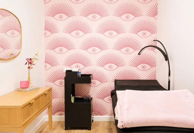 Room with pink eye-patterned wallpaper, black chair with pink blanket, wooden table, and black lamp.