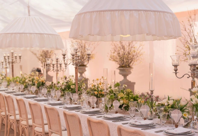 Elegant long dining table set with floral arrangements and large white pendant lamps in a tented venue.