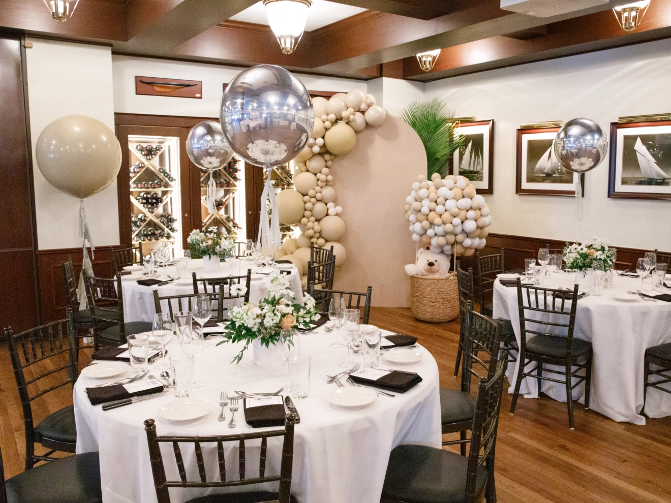 Elegant indoor dining setup with round tables, white tablecloths, floral centerpieces, and balloon decorations.