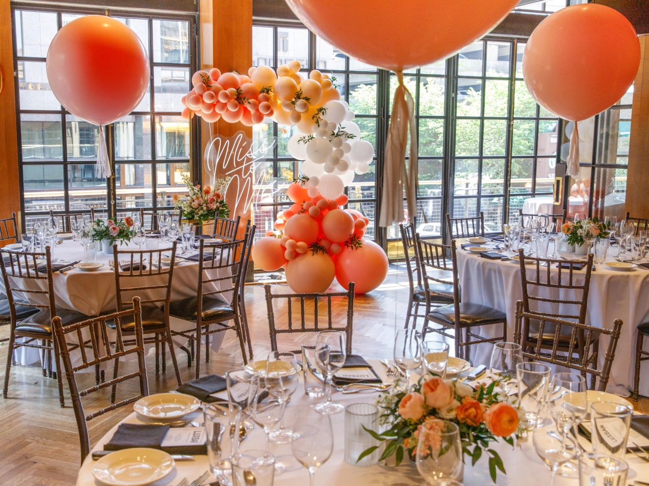 Elegant indoor event setup with orange and white balloon decorations, round tables, and floral centerpieces.