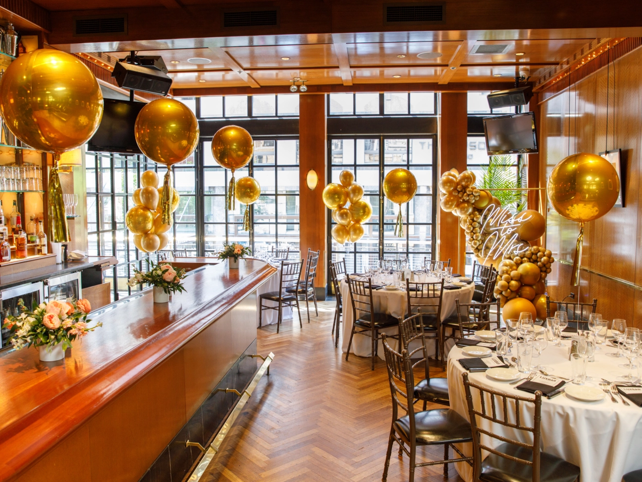 Indoor celebration setup with gold balloons, decorated tables, flowers, and a wooden bar counter.