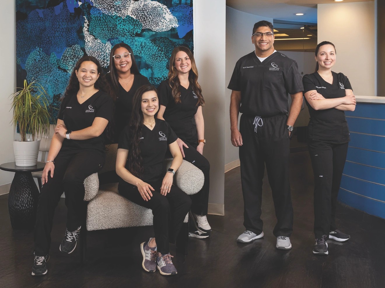 Group of six healthcare professionals in black scrubs posing indoors with a blue and white wall background.