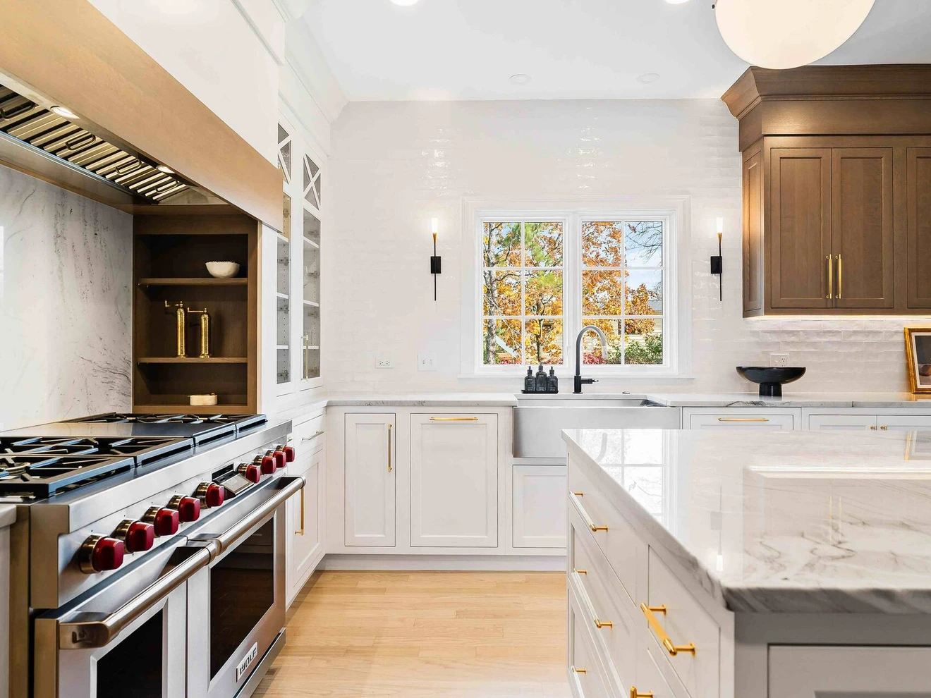 Modern kitchen with white and wood cabinetry, marble countertops, large stove, and window showing autumn trees.