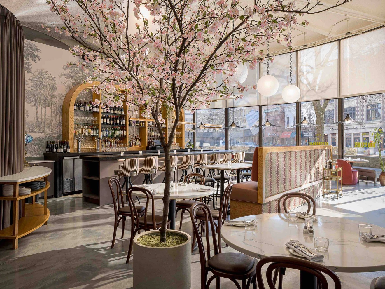 Bright, modern restaurant interior with marble tables, wooden chairs, large windows, and a flowering tree centerpiece.