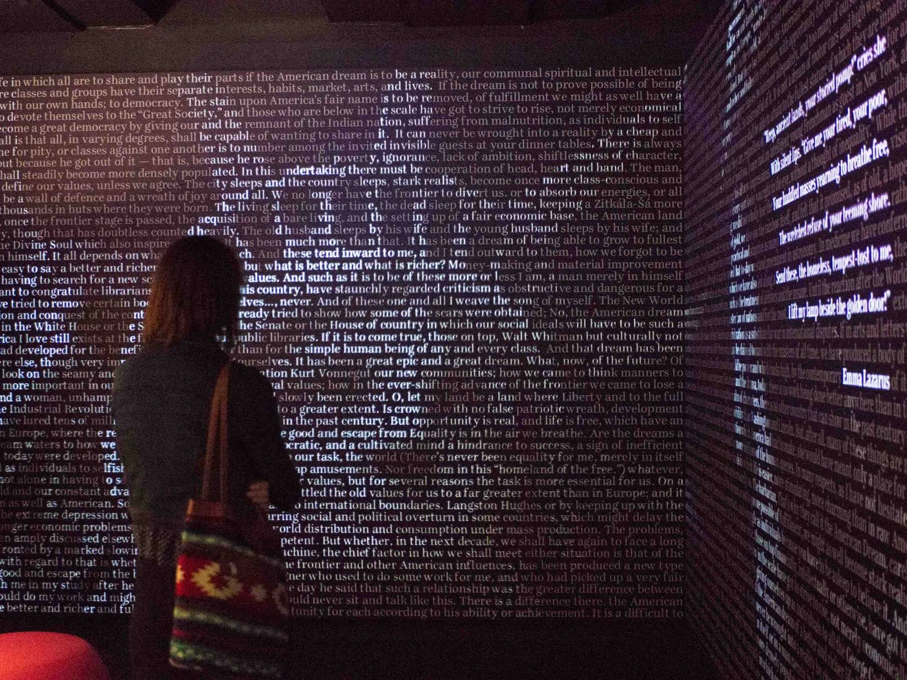 Person standing in front of a large wall covered in dense, illuminated text in a dark room.