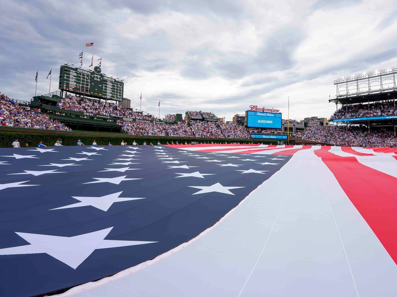 Large American flag displayed on the field of a crowded baseball stadium under a cloudy sky.
