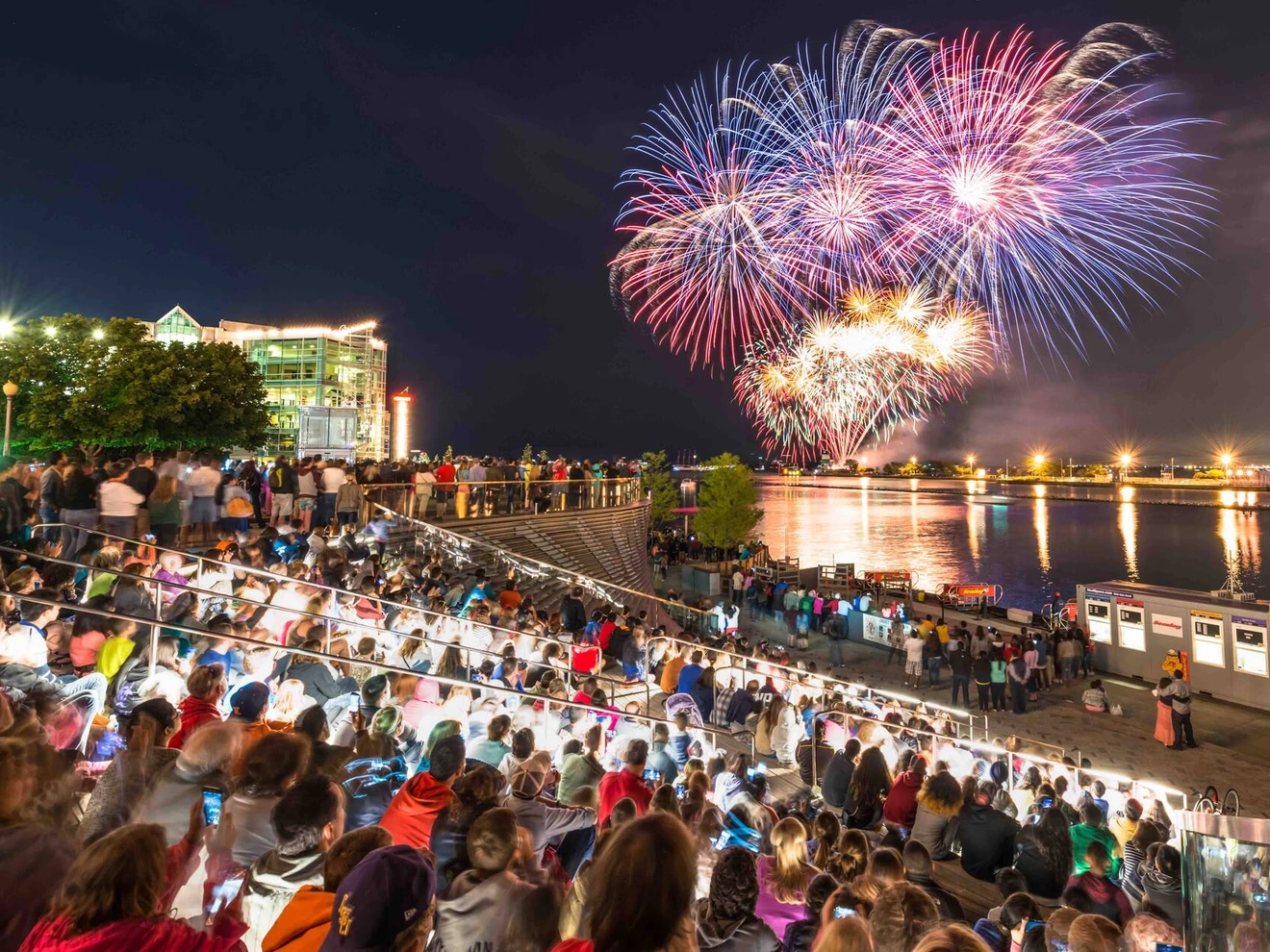 Crowd of people watching colorful fireworks over a river at night.