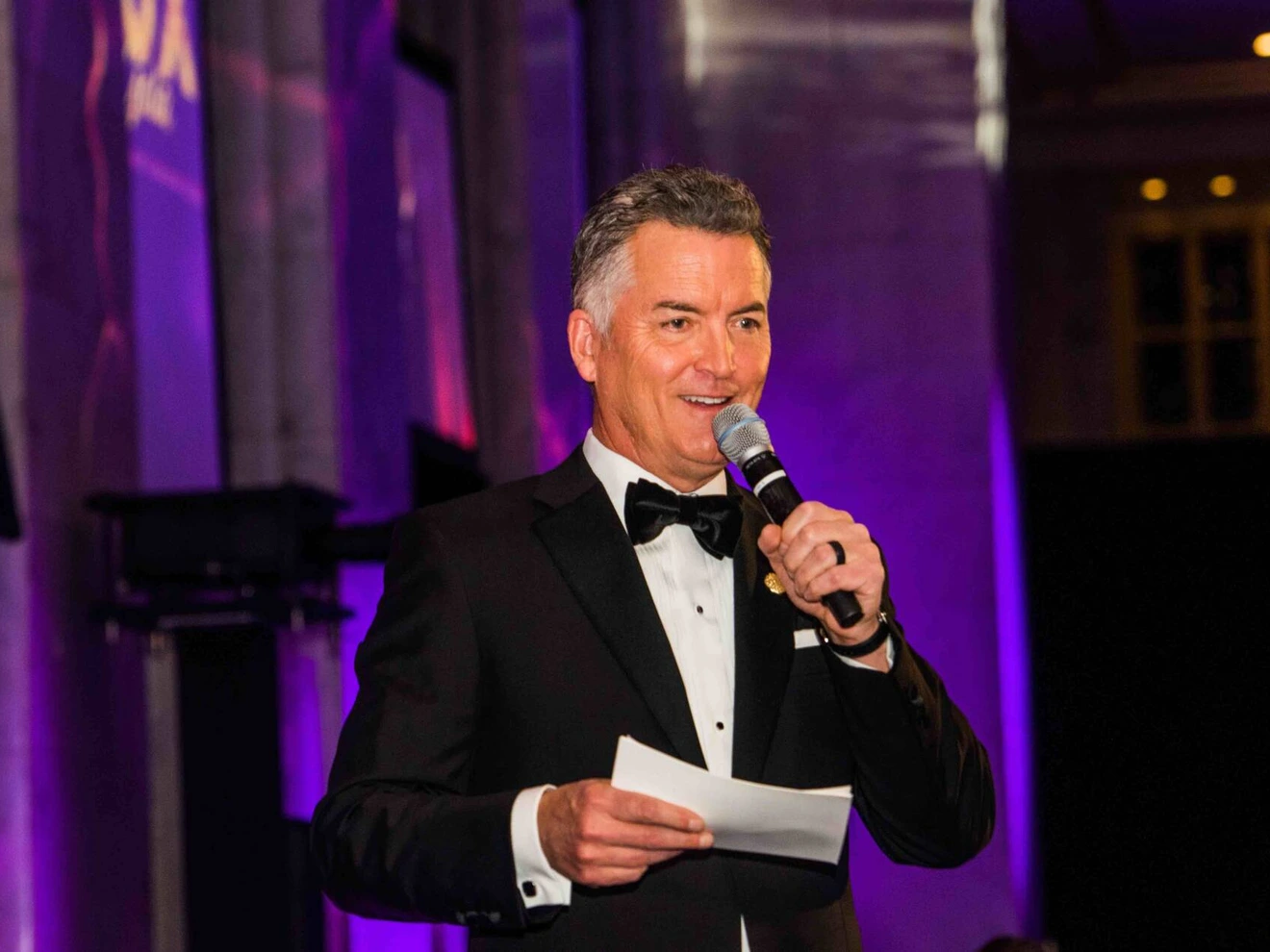 Man in tuxedo holding microphone and notes, speaking at formal indoor event.