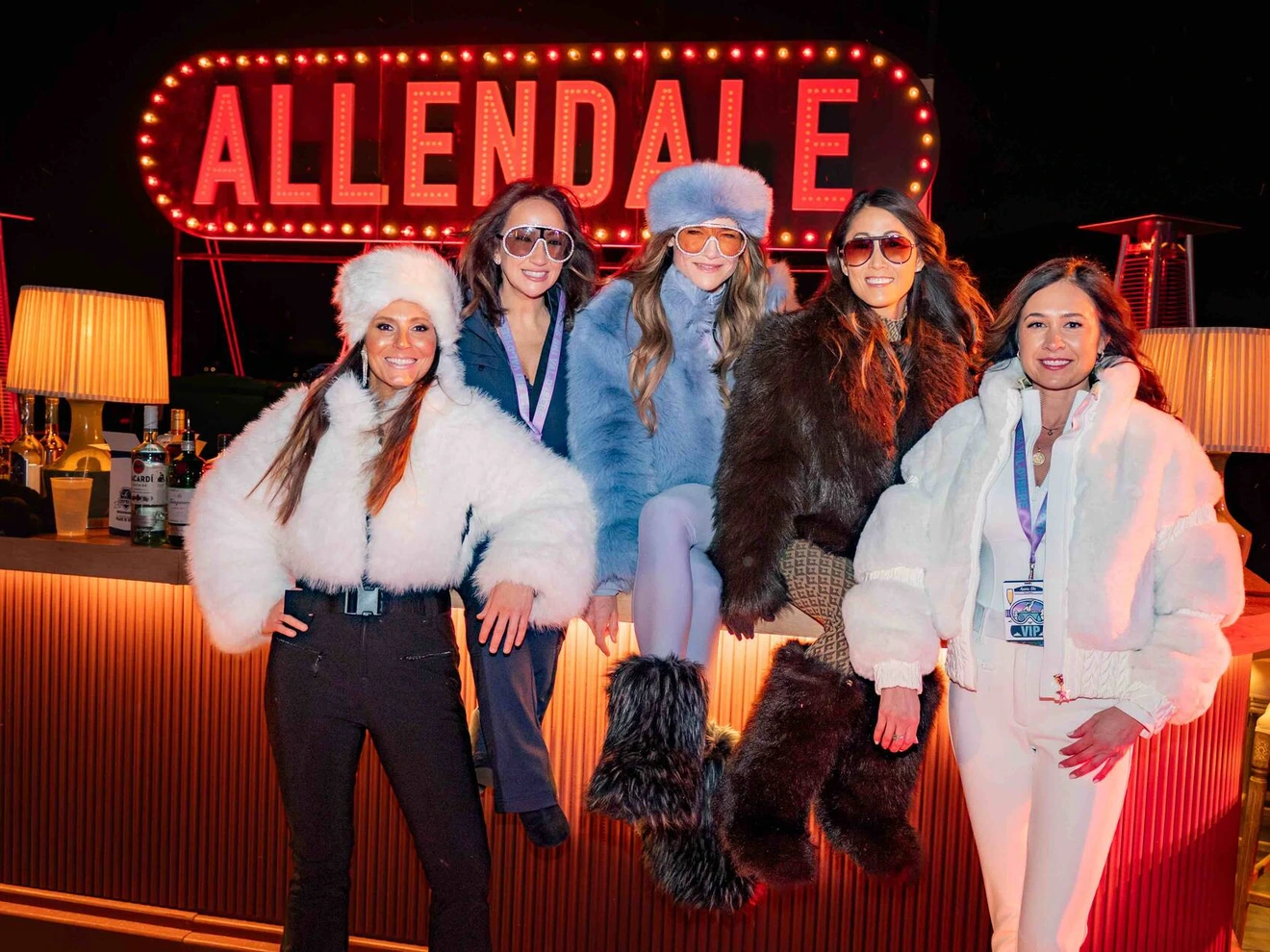 Five women wearing winter outfits posing at a bar with a bright ALLENDALE sign in the background.