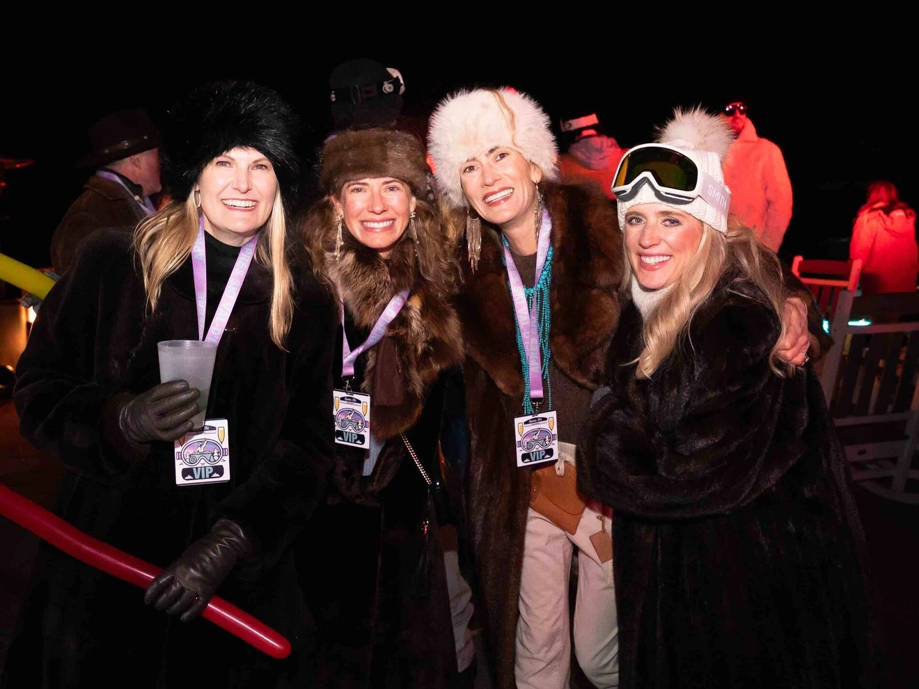 Four women dressed in winter fur coats and hats, smiling at a nighttime outdoor event.