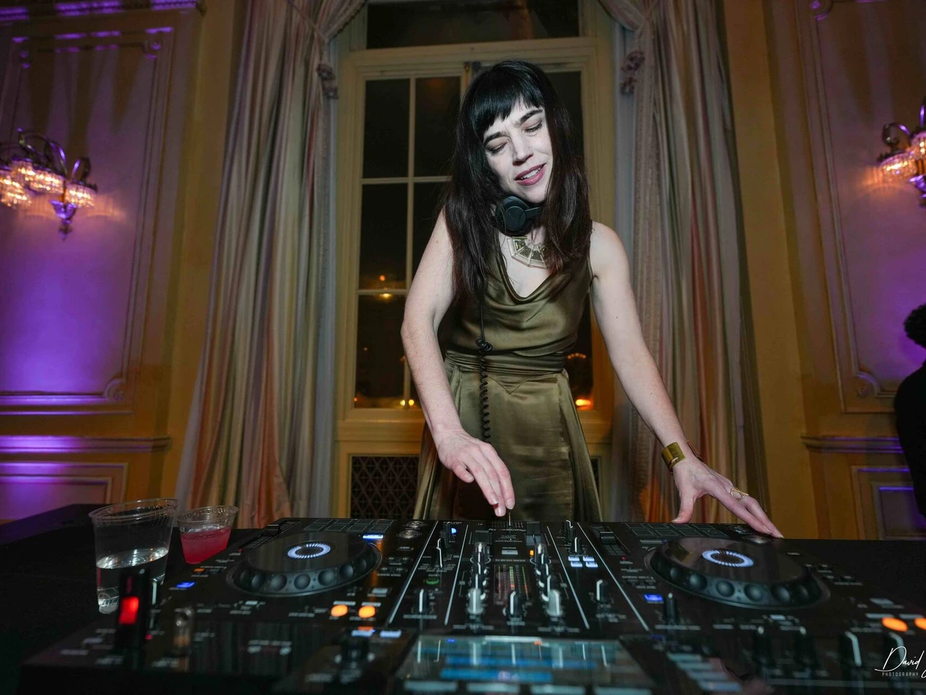Woman DJ wearing headphones, performing with mixer in an elegant room with purple lighting.
