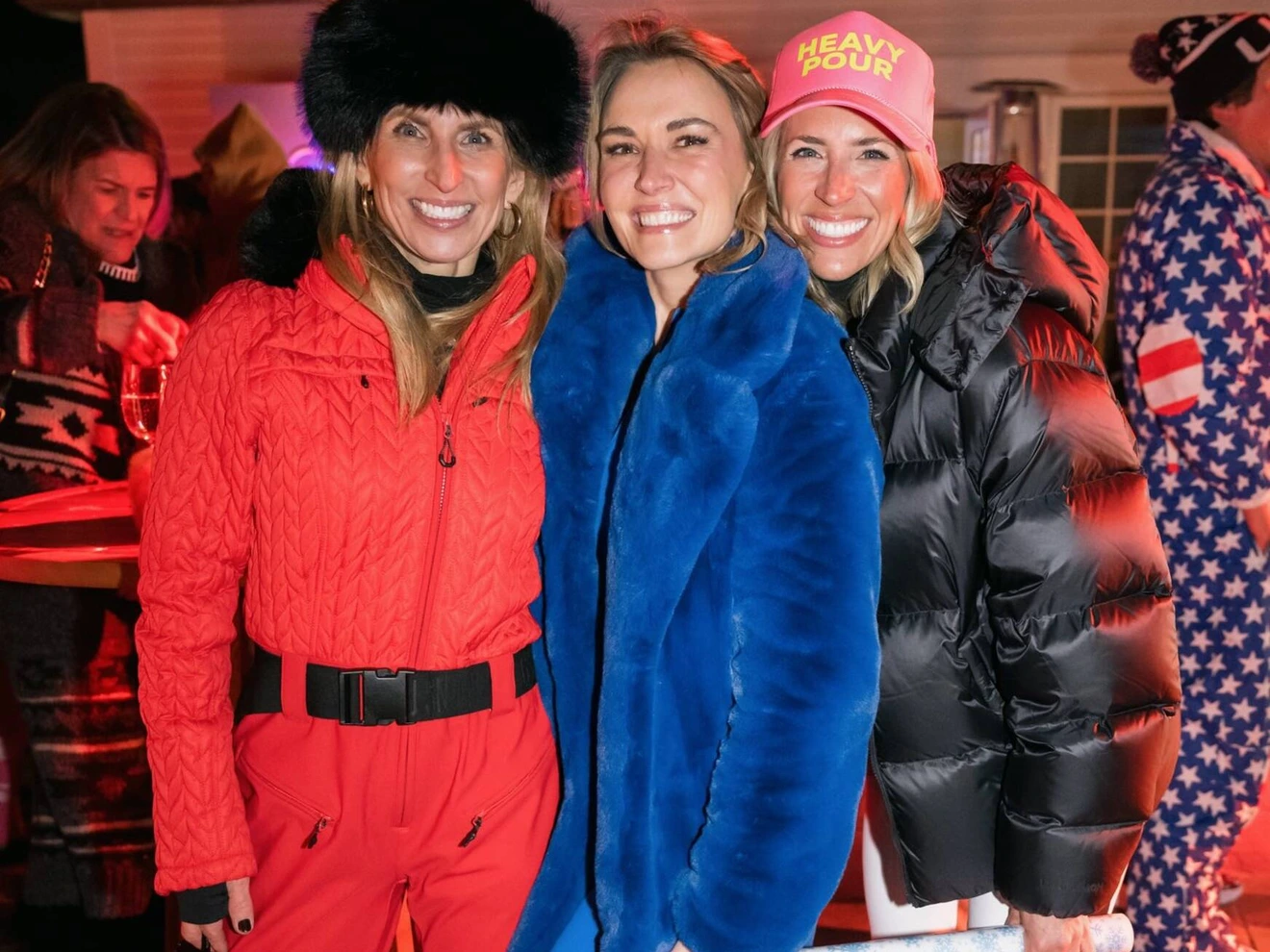 Three smiling women dressed in colorful winter clothing at an outdoor nighttime event.