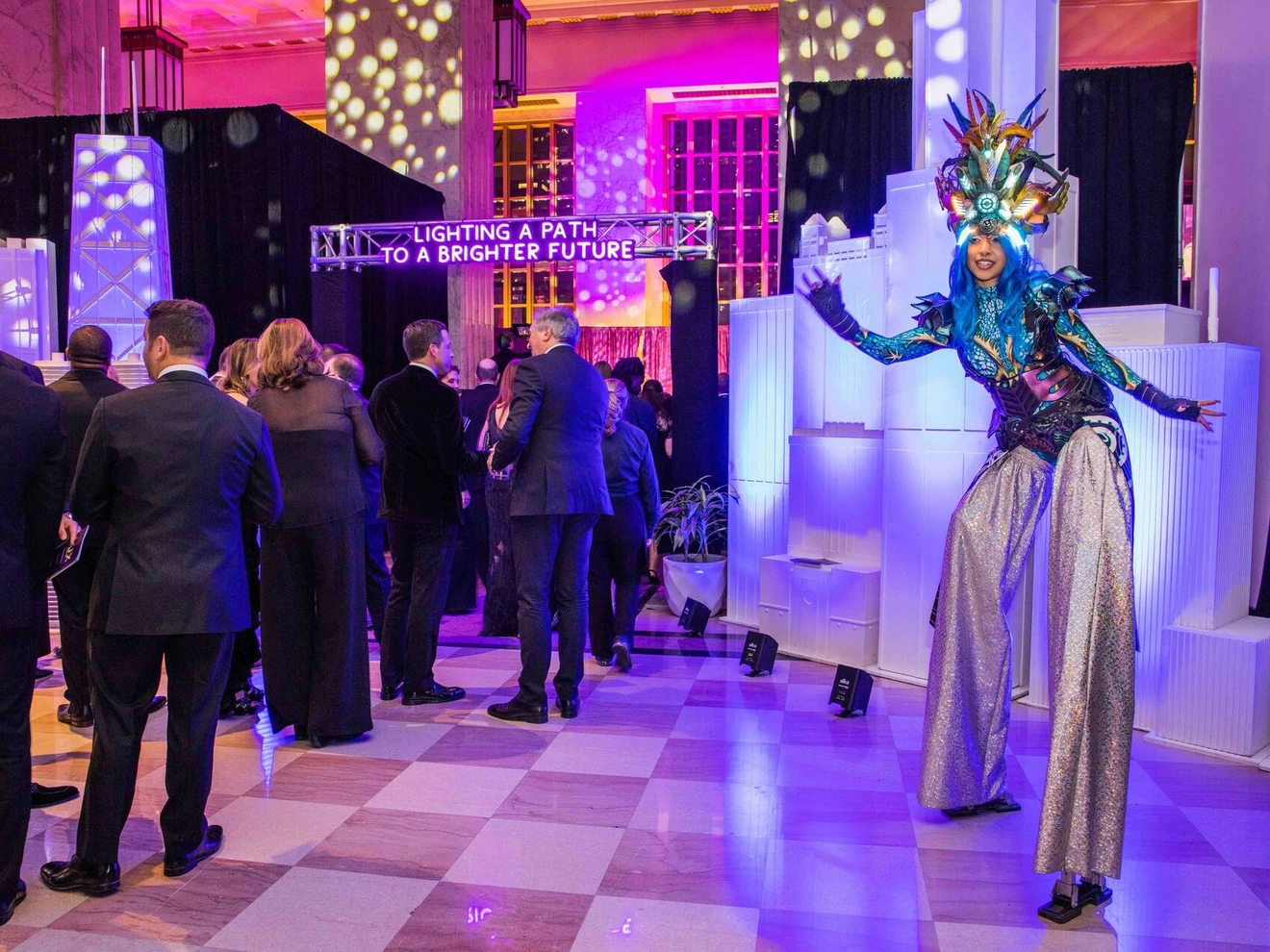 People in formal attire at an event with a performer on stilts in a colorful costume.