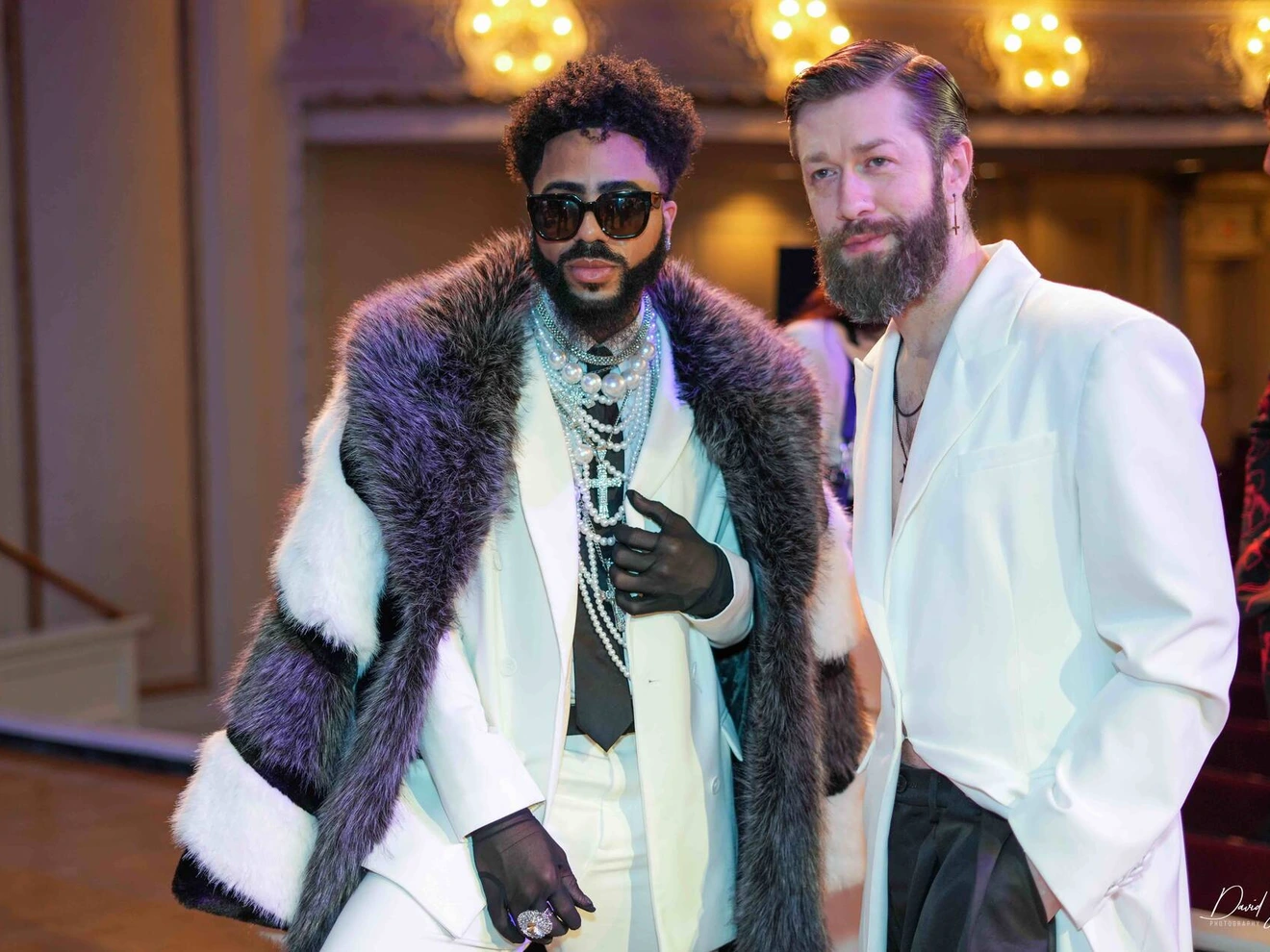 Two stylishly dressed men, one wearing sunglasses and a fur-trimmed coat, posing indoors at an event.