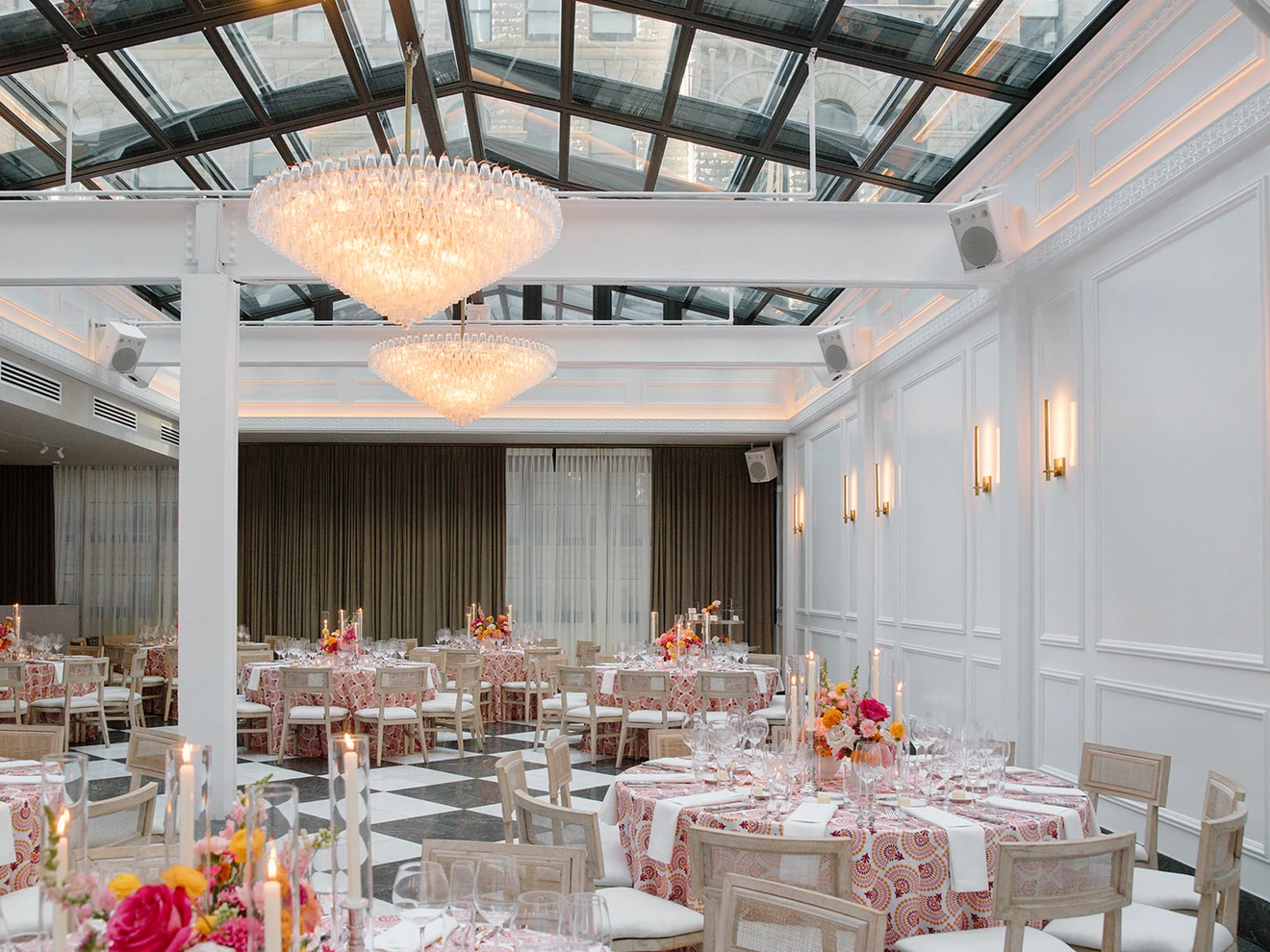 Elegant banquet hall with round tables, floral centerpieces, chandeliers, and checkered floor under a glass ceiling.