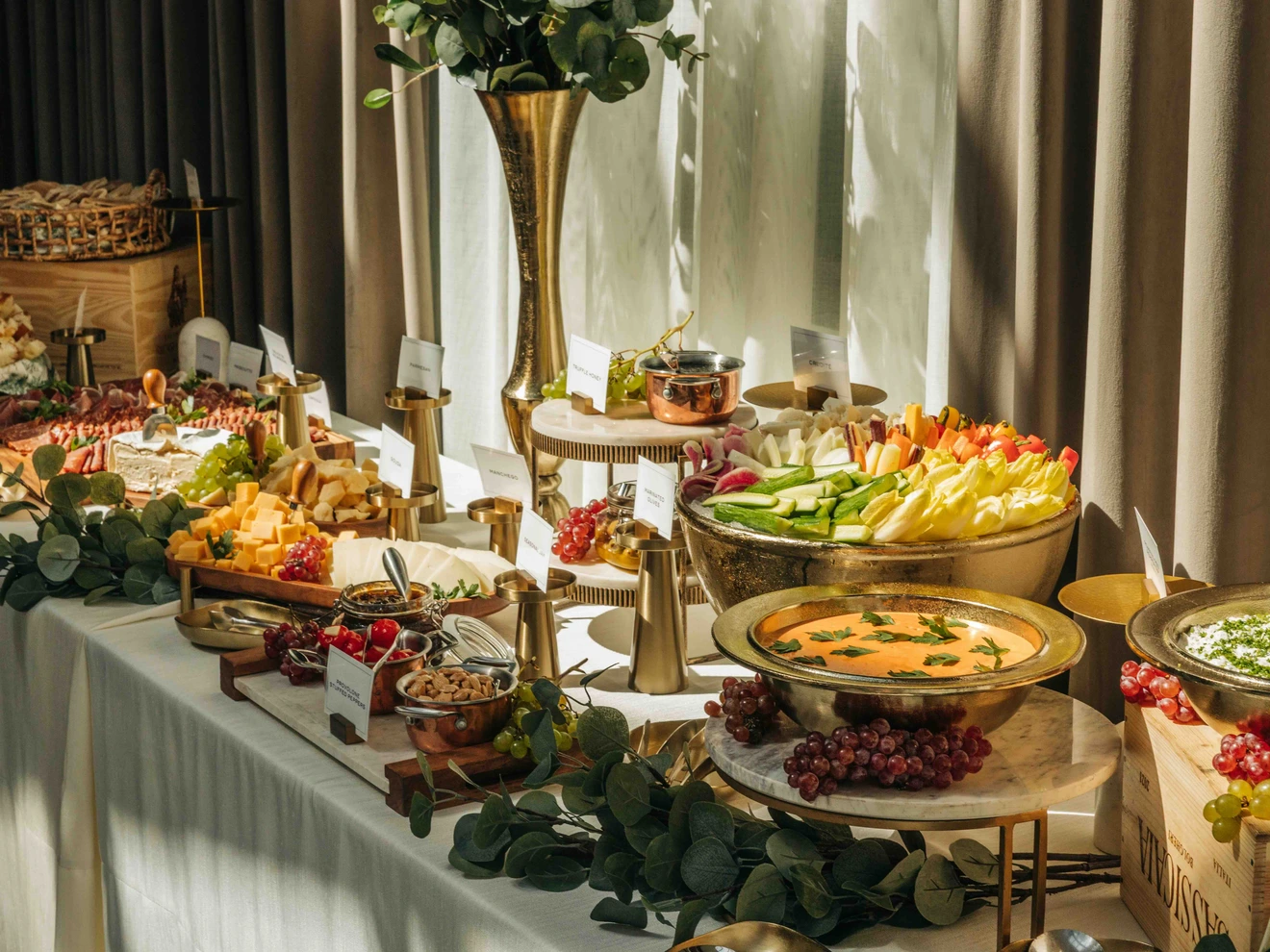 Buffet table with assorted vegetables, dips, cheeses, and fruits arranged with decorative greenery.
