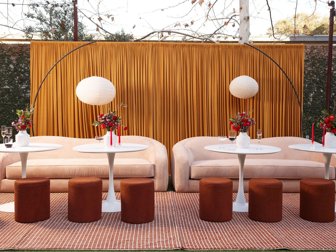 Outdoor seating area with beige sofas, round white tables, brown stools, red floral centerpieces, and hanging round lamps.