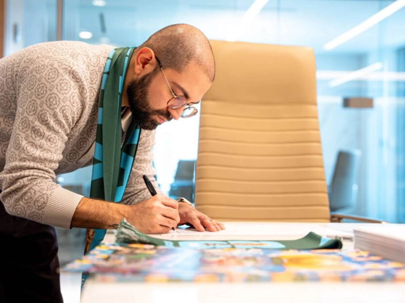 Man with glasses and scarf writing on paper at a desk in a modern office.