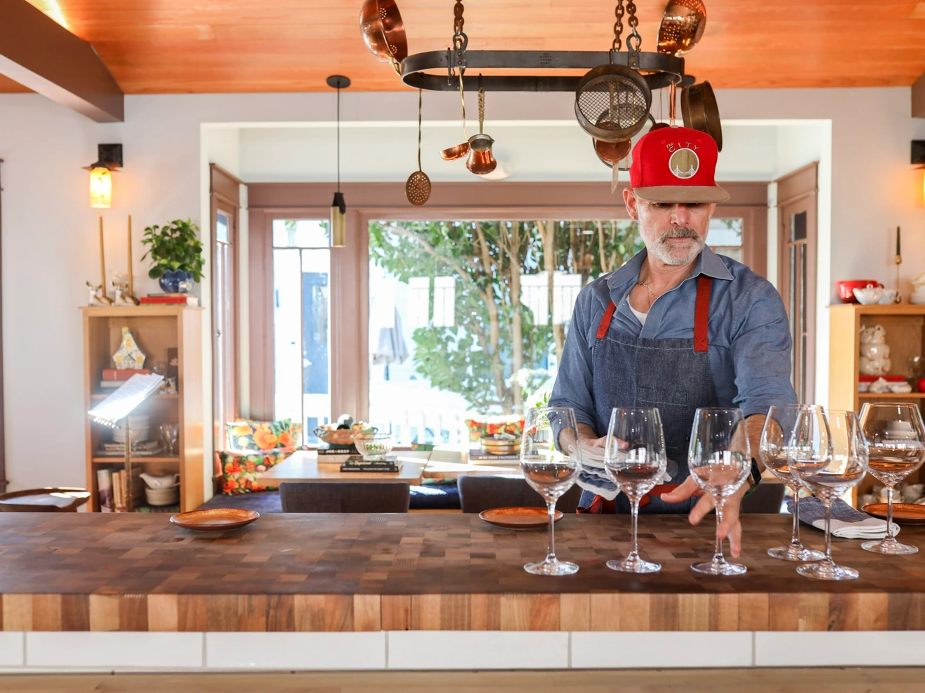 Man in red cap and apron setting wine glasses on wooden kitchen counter.