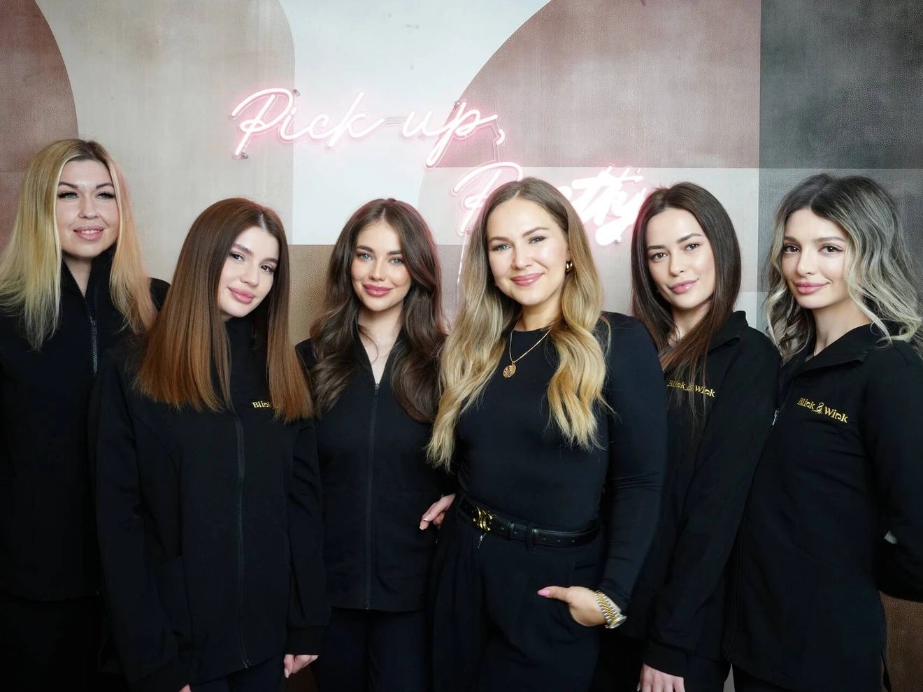 Group of six women posing indoors in front of a neon sign reading Pick up, Pretty.