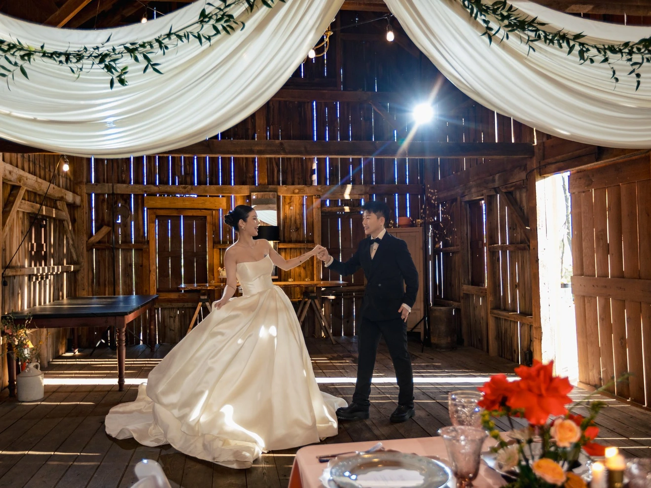Bride and groom dancing in a rustic barn with draped white fabric and floral decorations.