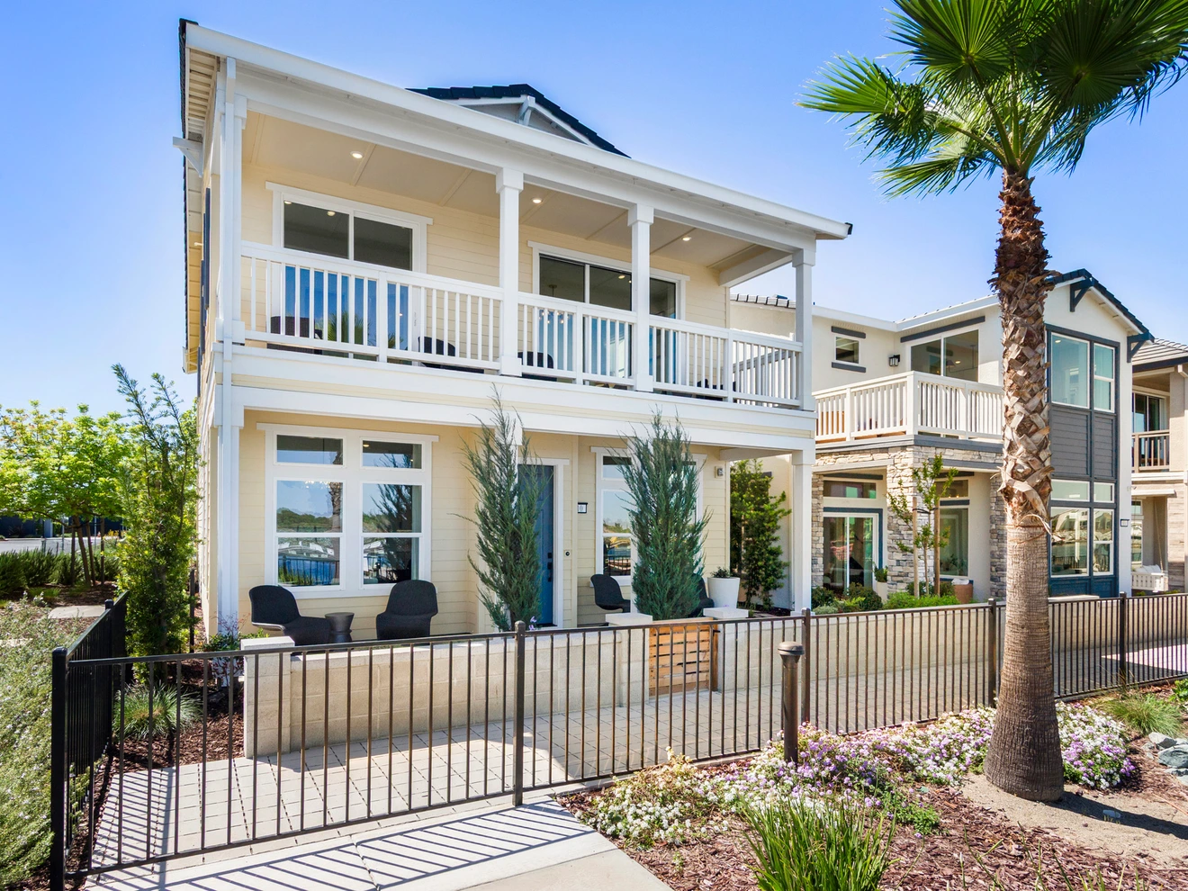 Two-story modern house with balconies, patio seating, black metal fence, and palm tree in sunny residential area.