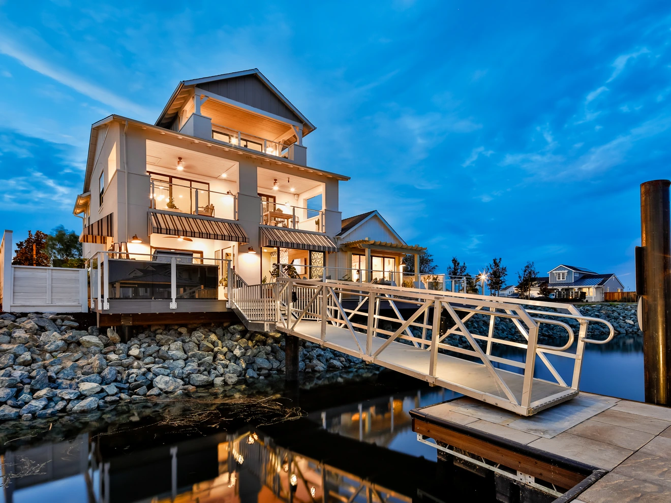 Modern three-story waterfront house with dock and gangway at dusk, calm water and blue sky.