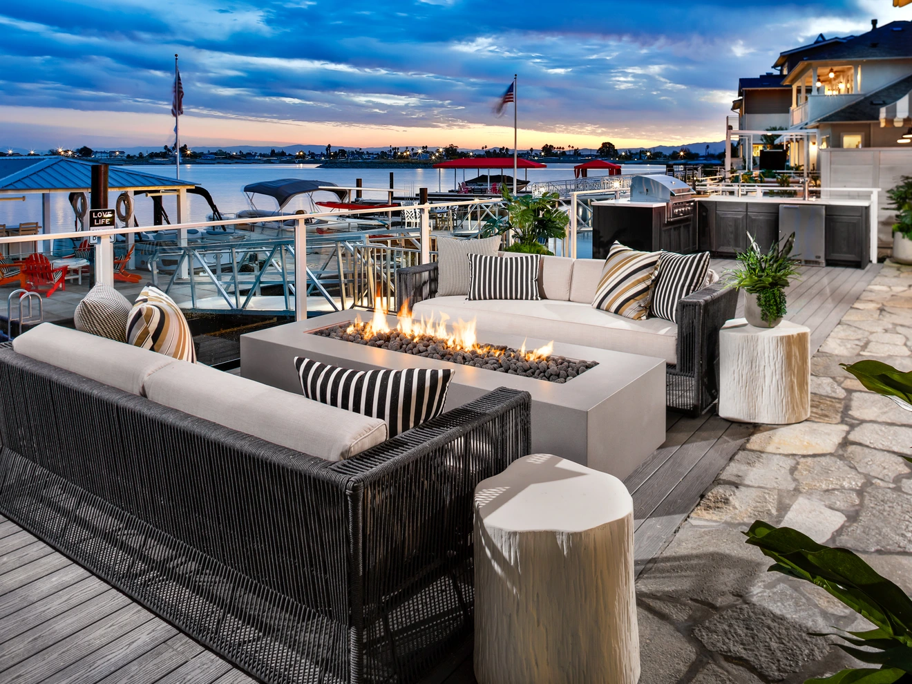 Outdoor seating area with modern sofas, fire pit, and waterfront view at sunset.