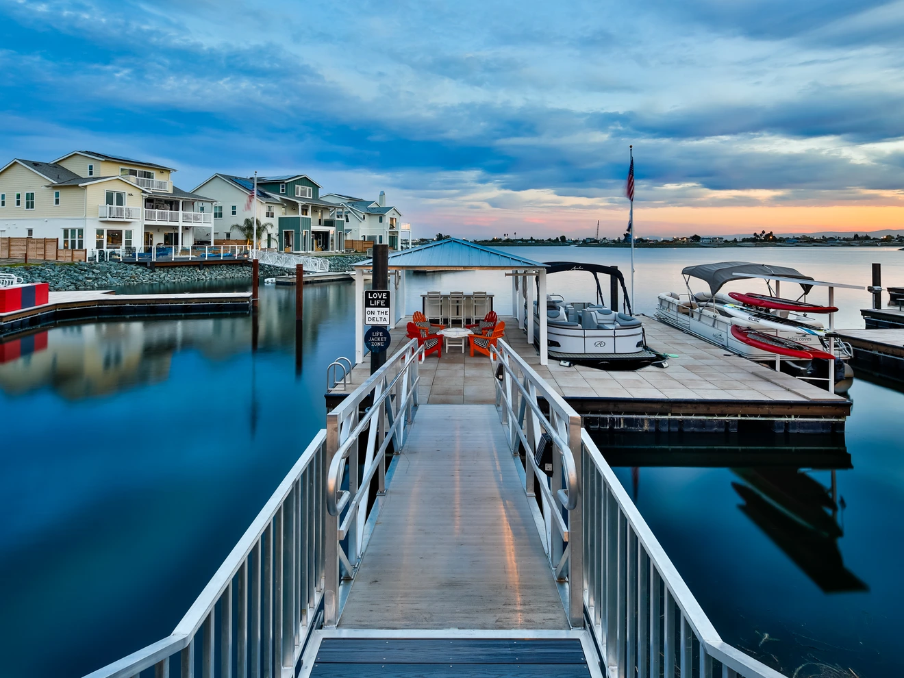 Dock with boats and houses along calm waterfront at sunset under a partly cloudy sky.