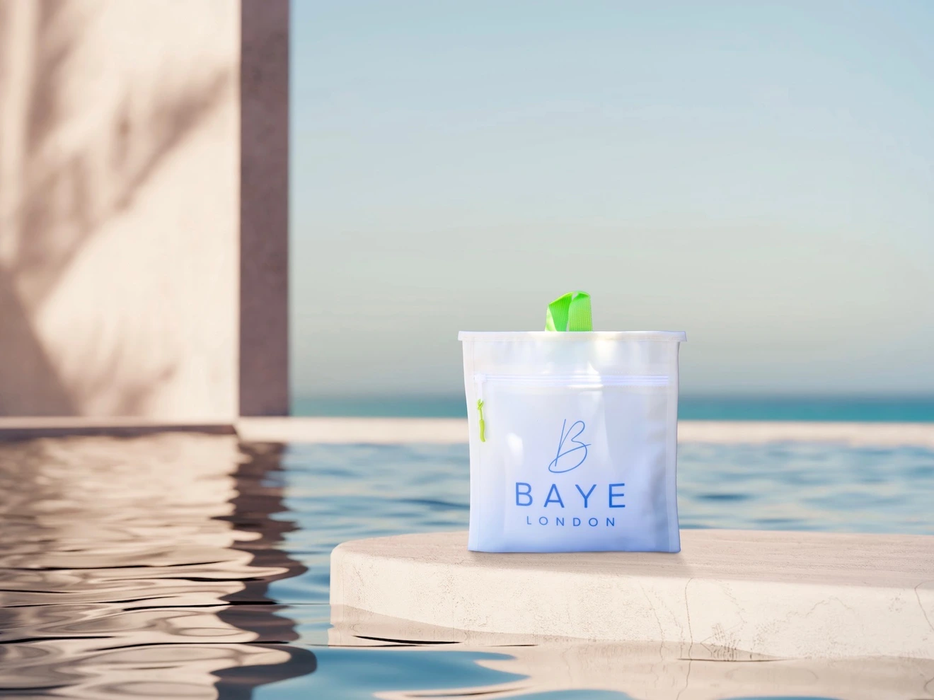 Transparent bag with Baye London logo, placed on a ledge near a calm pool and clear sky.
