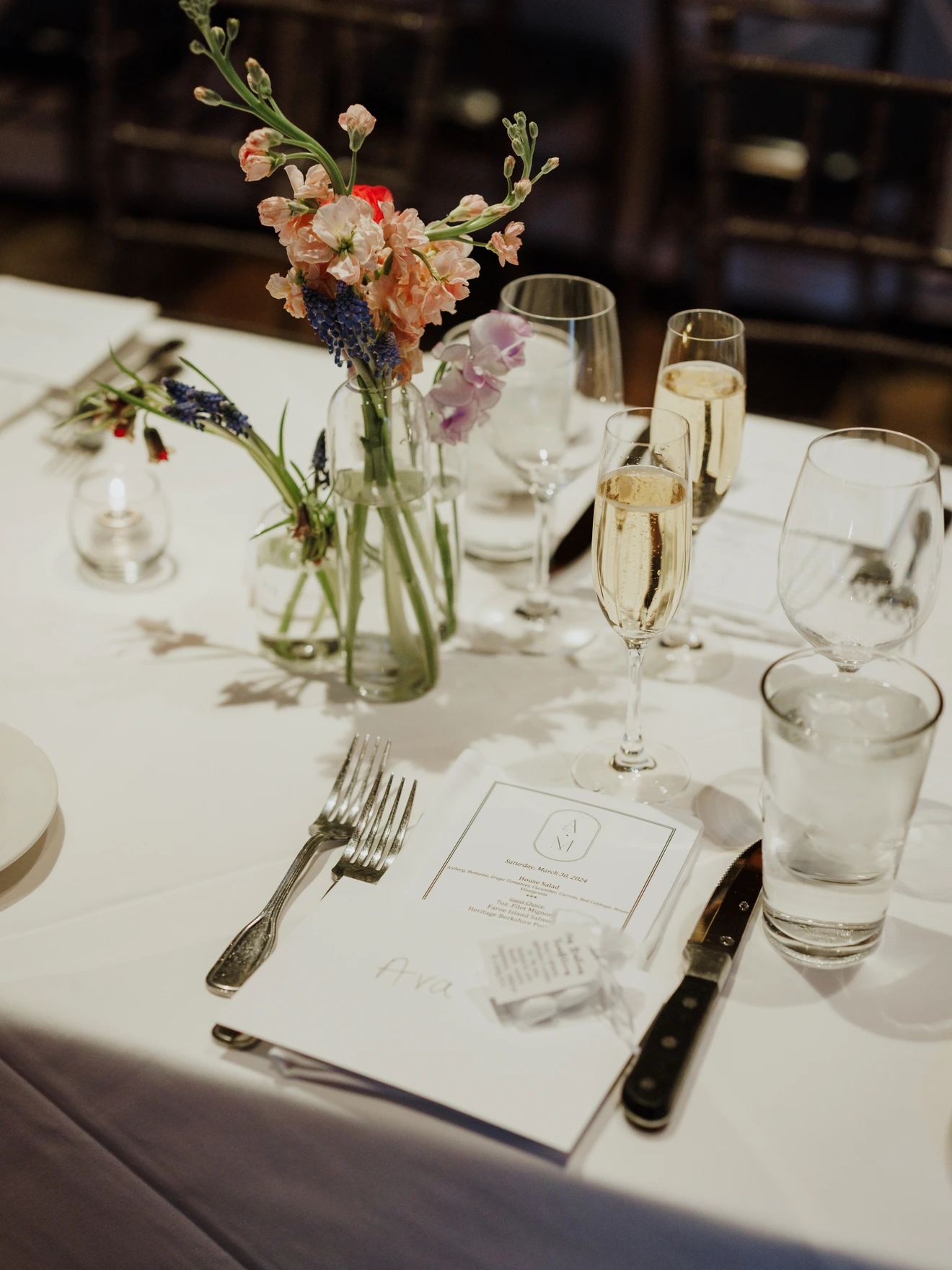 Table setting with flowers in glass vases, champagne glasses, water glass, cutlery, and a menu on a white tablecloth.