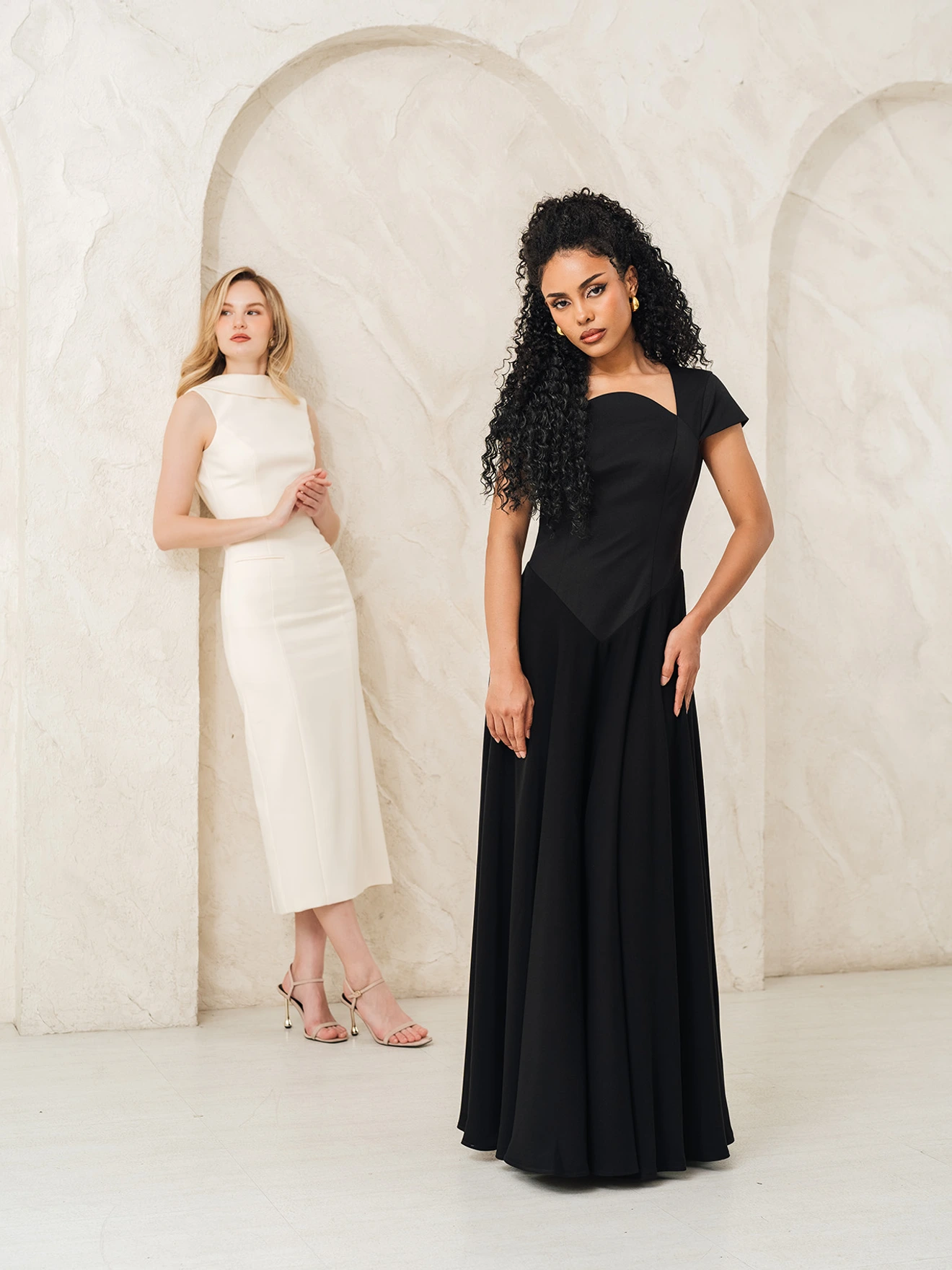 Two women modeling dresses. The woman farther in the back is wearing a white dress. The woman in the foreground is wearing a black dress.