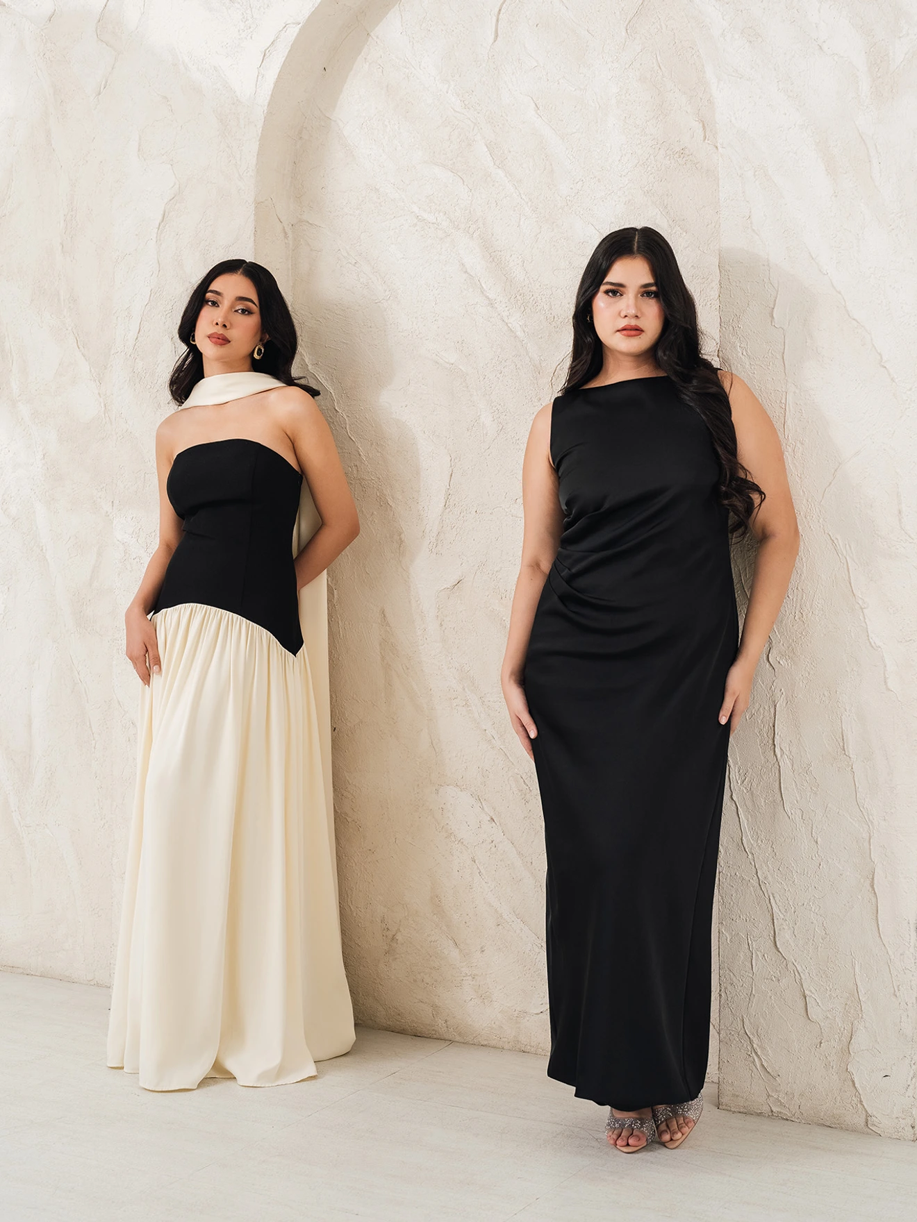 Two women facing forward, posing. The woman on the left is wearing a black and white dress, the woman on the right is wearing a black dress.