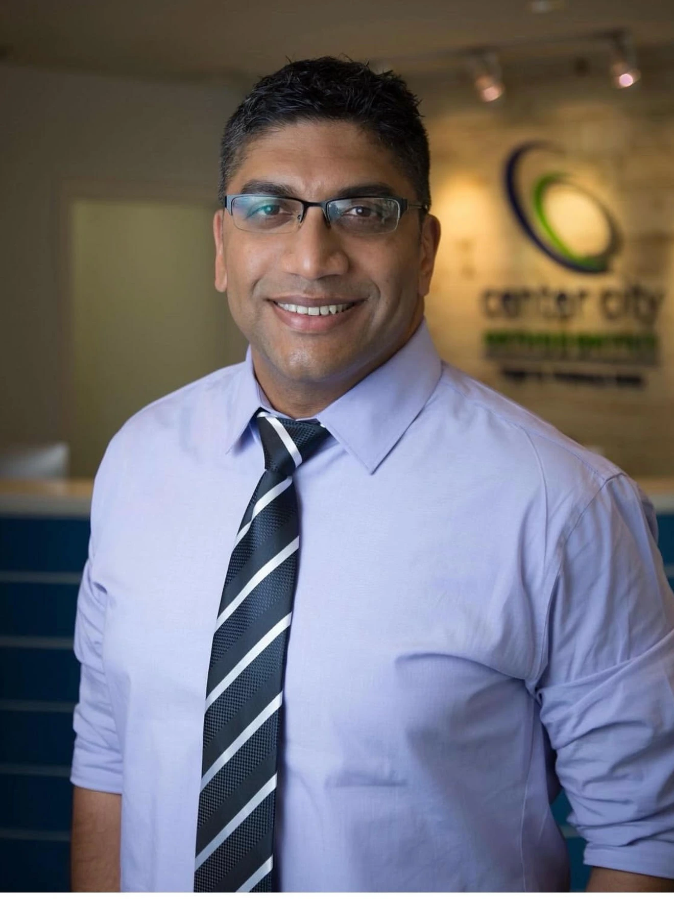 Smiling man wearing glasses, light purple shirt, and striped tie in a professional office setting.