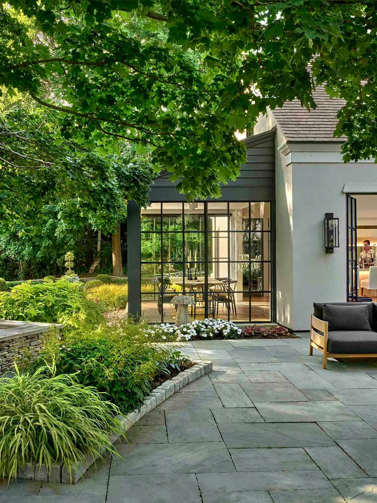 Modern outdoor patio with stone flooring, wooden furniture, and lush green garden surrounding a white house.