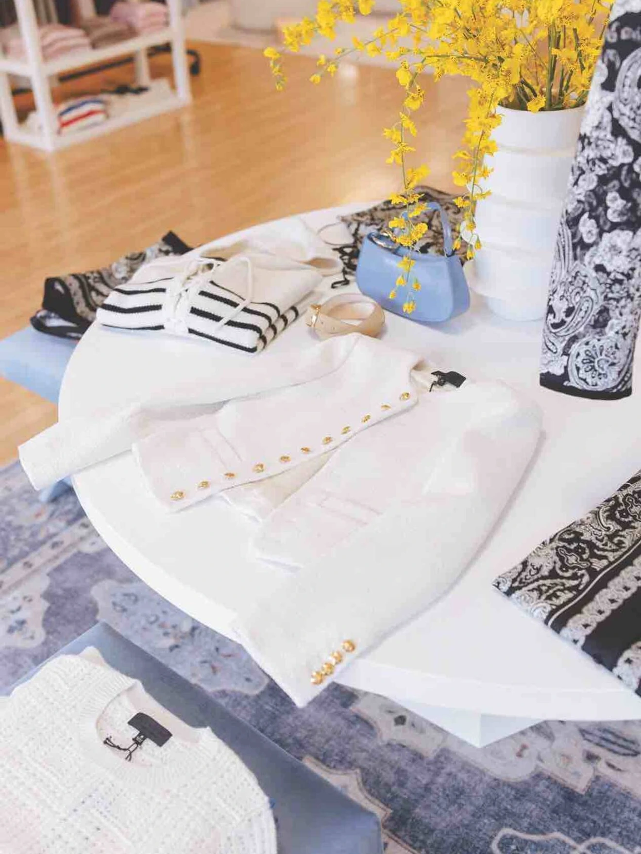 Round white table displaying folded white and striped clothing, a blue handbag, yellow flowers in a vase.