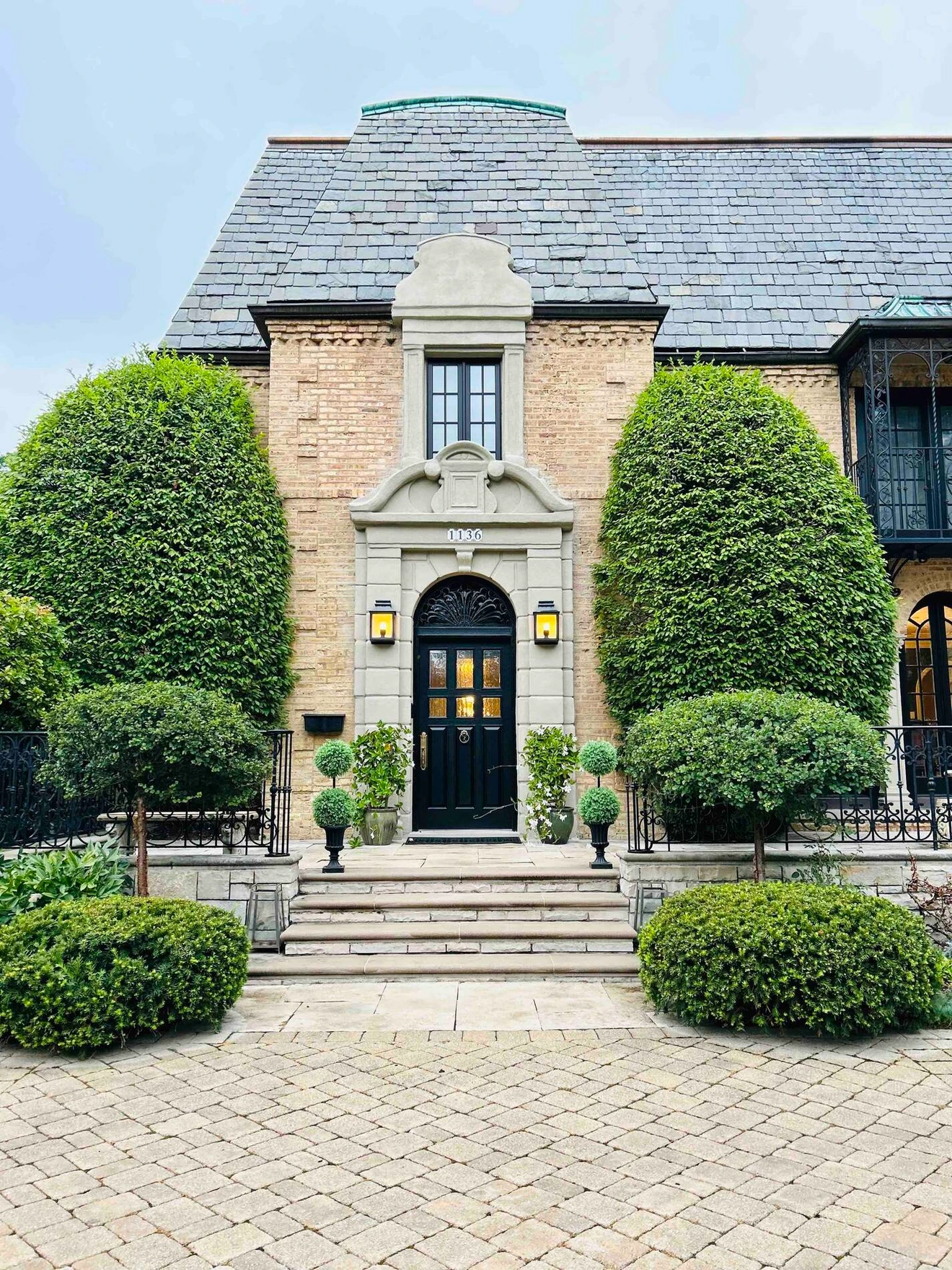 Elegant brick house entrance with black door, symmetrical topiary trees, and stone steps.