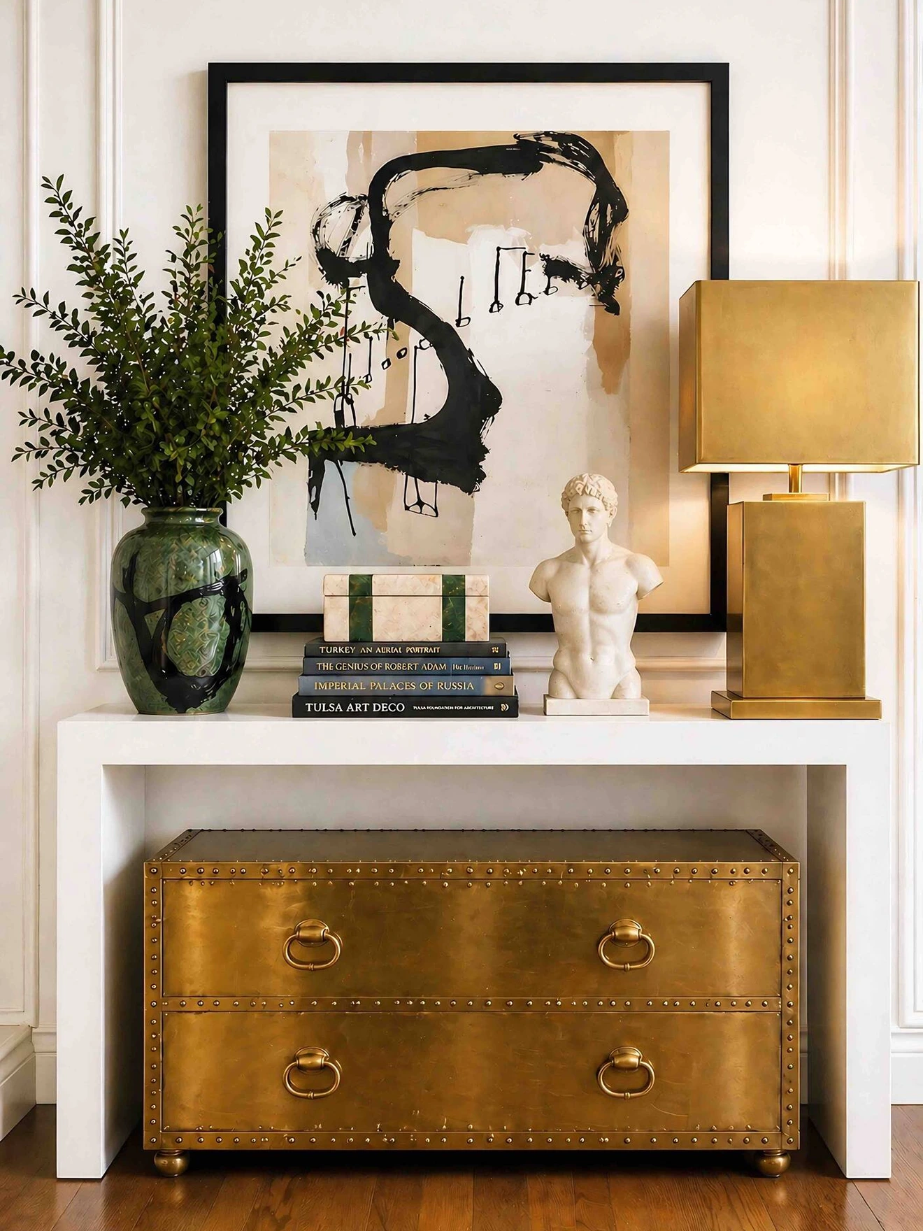 Console table with abstract art, green vase with plant, white bust statue, stacked books, gold lamp, and large gold drawer chest.