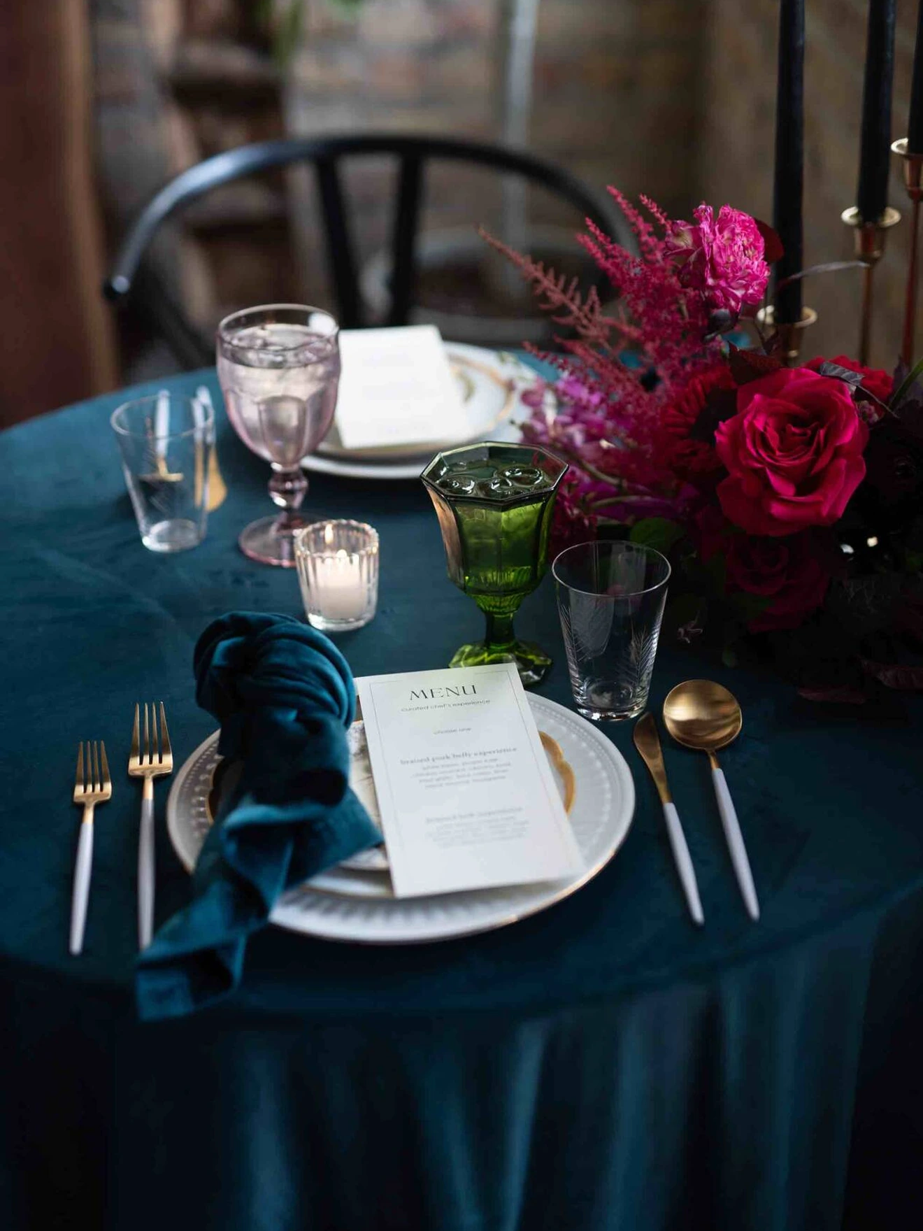 Elegant table setting with dark teal tablecloth, gold utensils, crystal glasses, and vibrant pink floral centerpiece.
