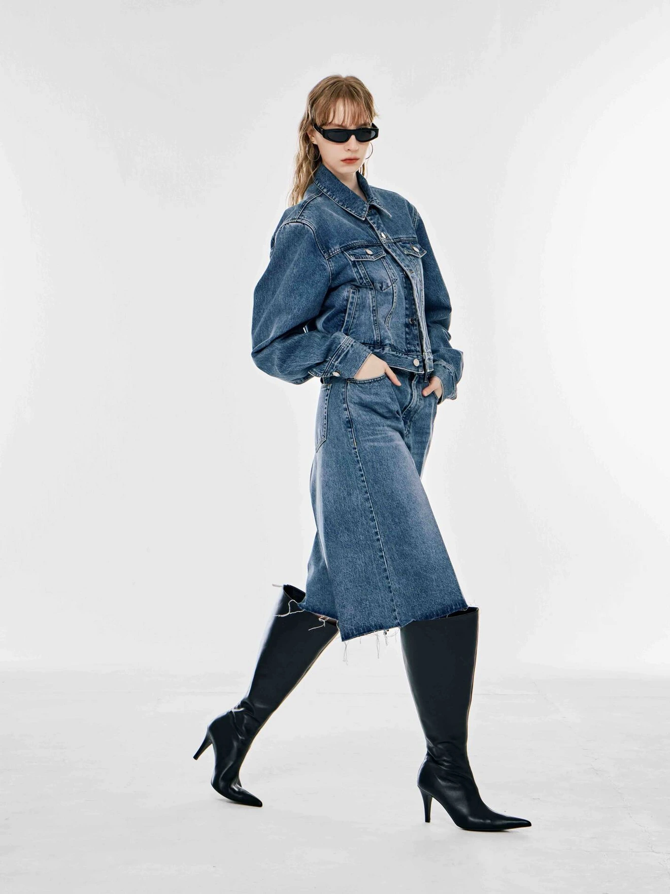 Woman in denim jacket and skirt, wearing black heeled boots and sunglasses, posing against a plain background.