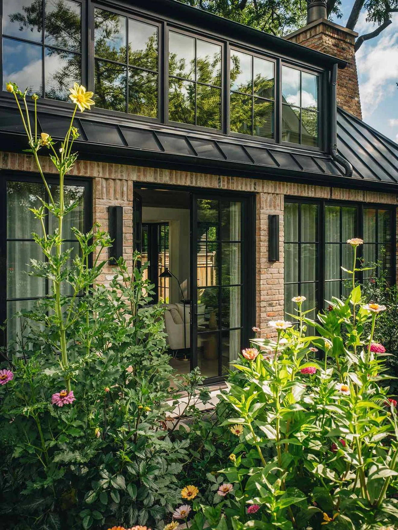 Modern brick house with large black-framed windows surrounded by tall, colorful garden flowers.