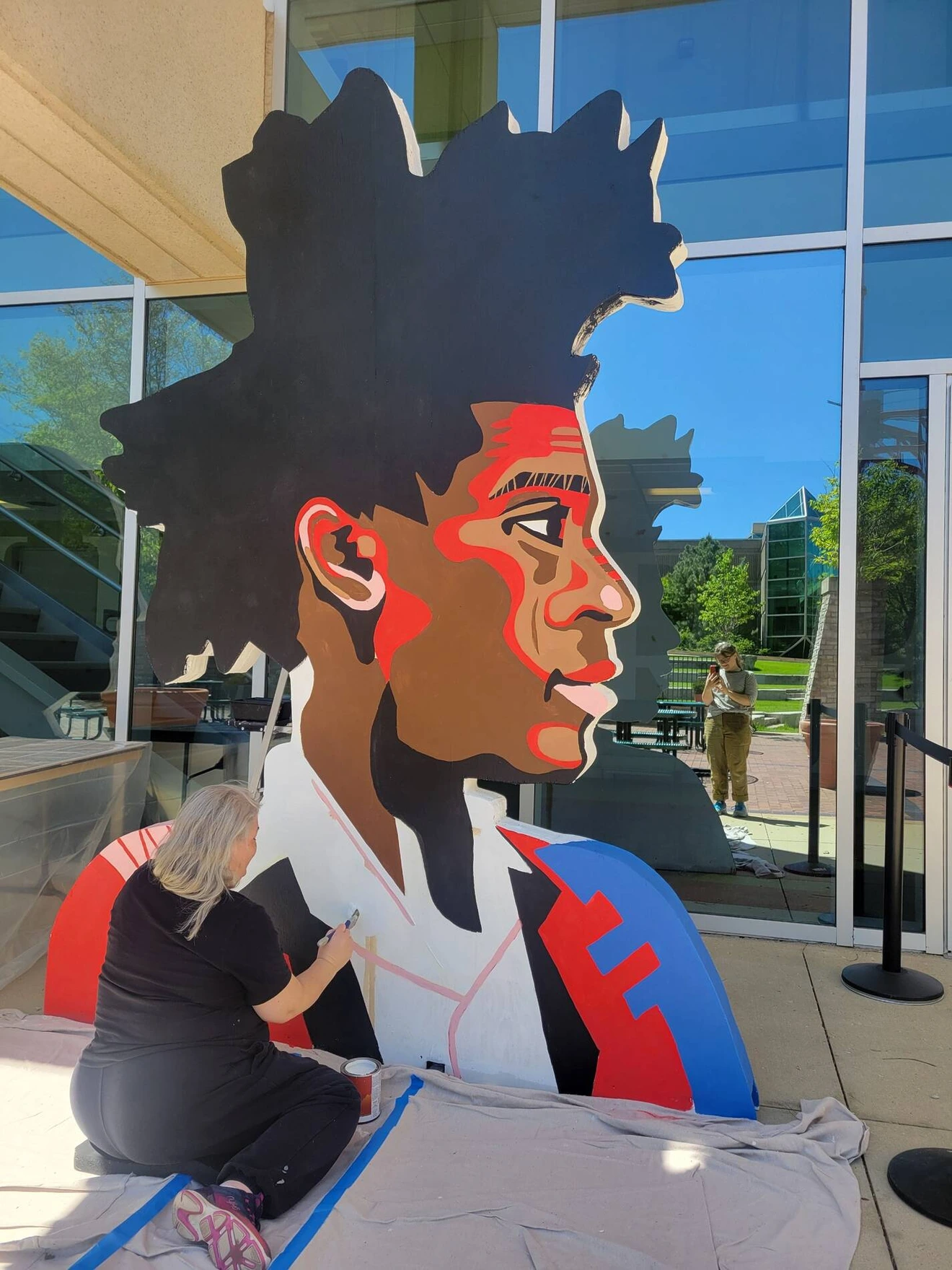 Artist painting large colorful portrait sculpture of a man with spiky hair outside a modern building.