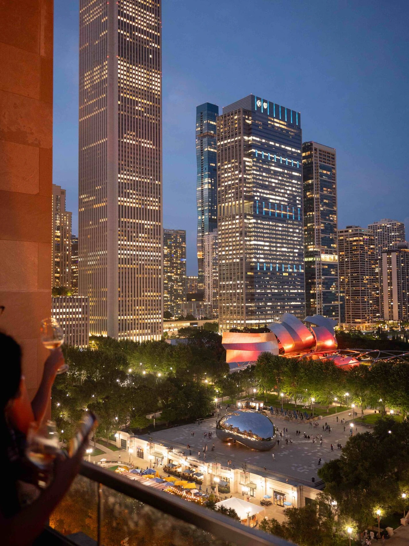 City skyline at dusk with illuminated skyscrapers and a park with people and unique architecture.