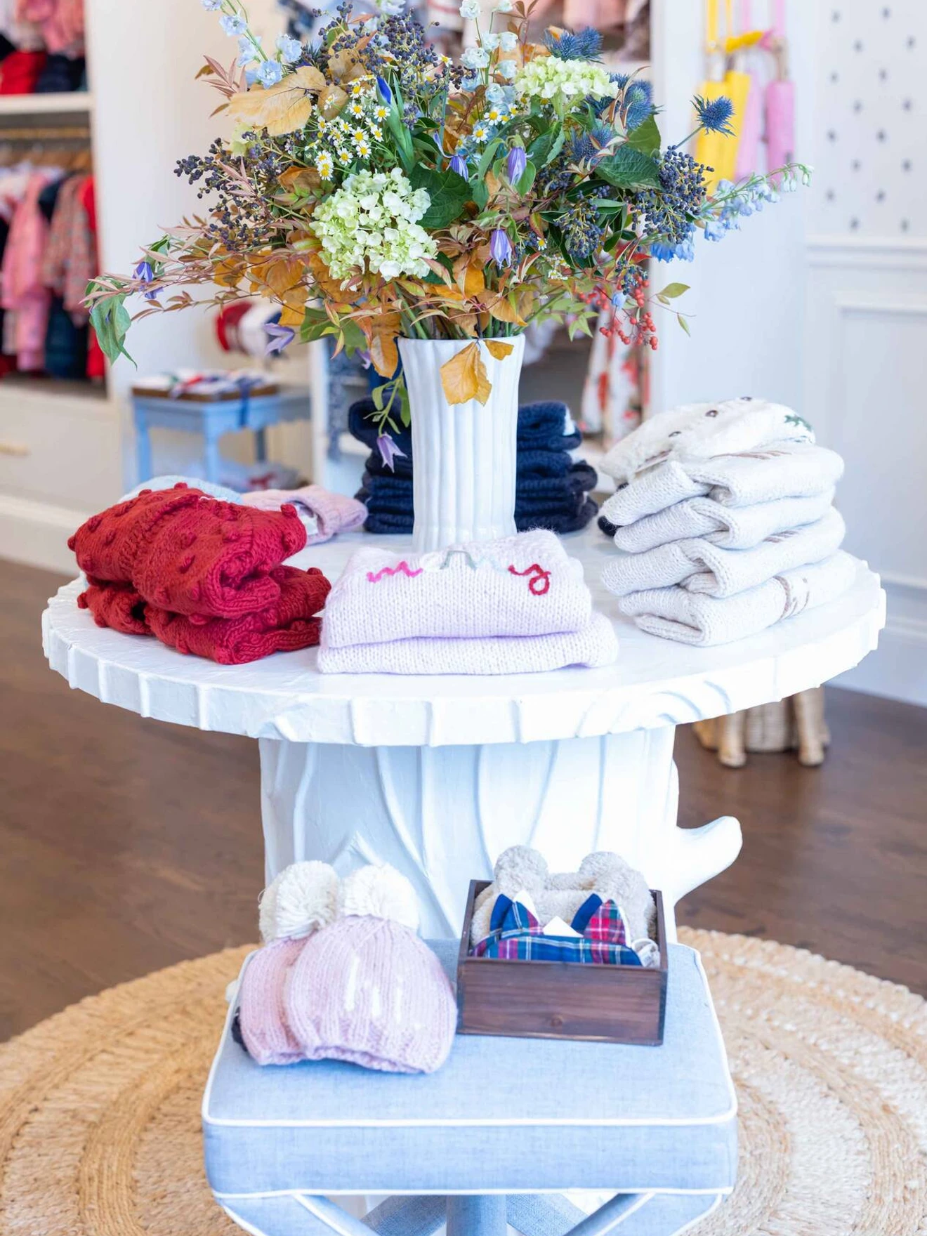 Round white table with folded clothes, a floral arrangement, and baby accessories in a boutique setting.