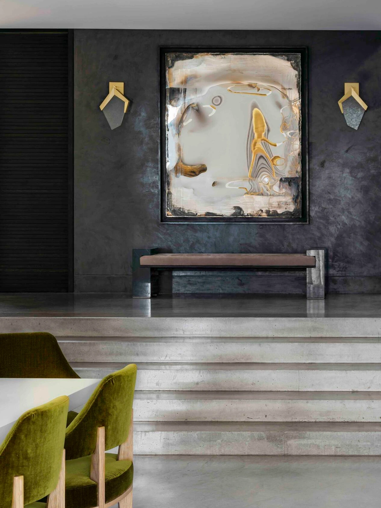 Modern interior with abstract painting, concrete stairs, green velvet chairs, and dark textured walls with sconces.