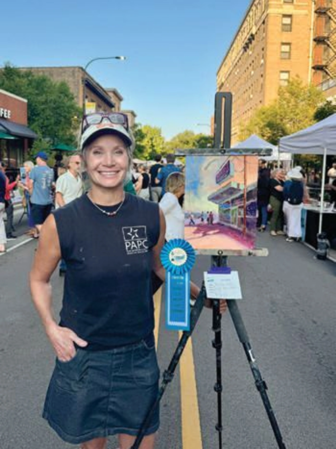 Woman smiling next to easel with painting and blue ribbon at outdoor art event on city street.