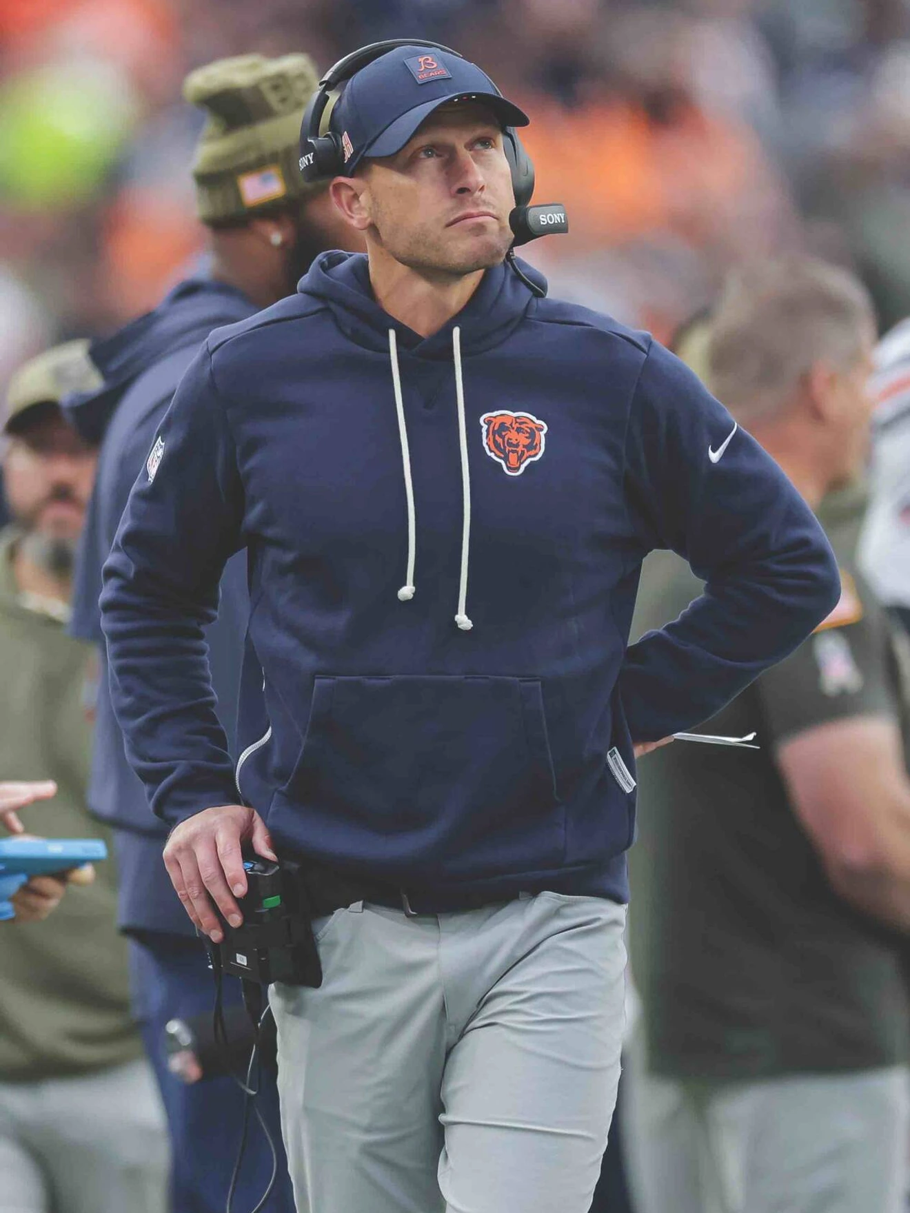 Coach wearing Chicago Bears hoodie and headset standing on football field during game.