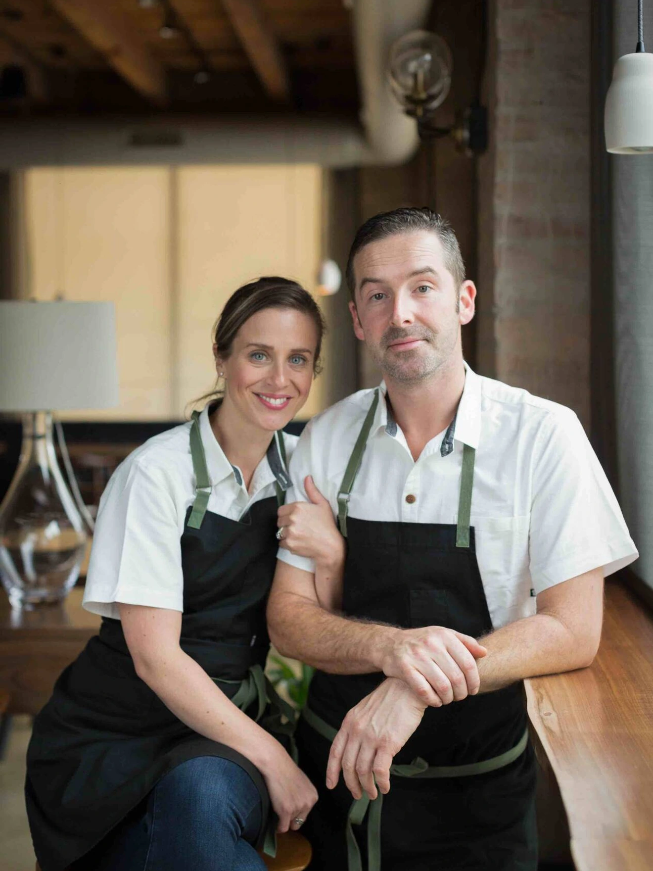 Portrait of a smiling man and woman wearing white shirts and black aprons in a cozy indoor setting.