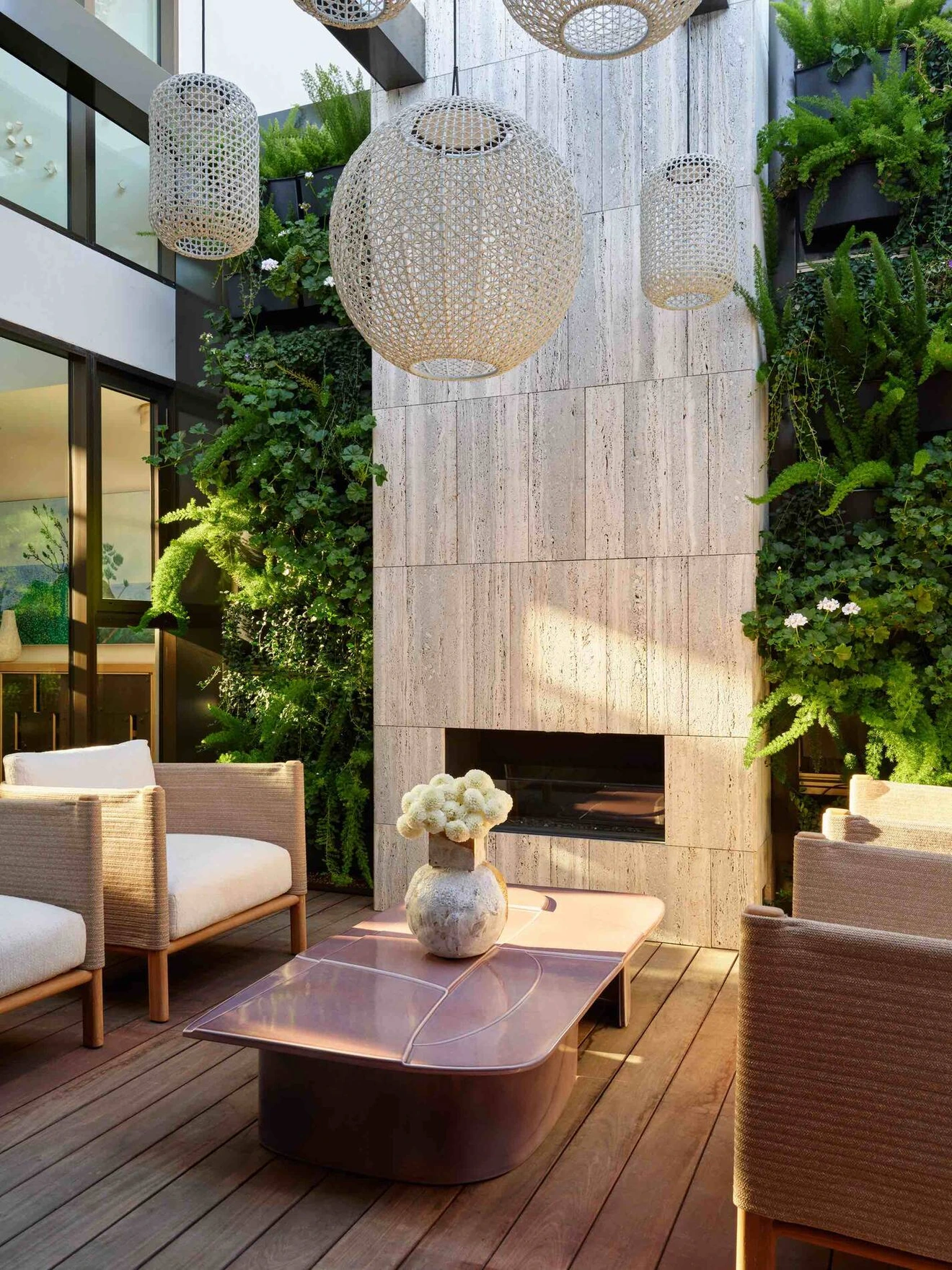 Modern outdoor seating area with stone fireplace, hanging lanterns, greenery, and wooden flooring.