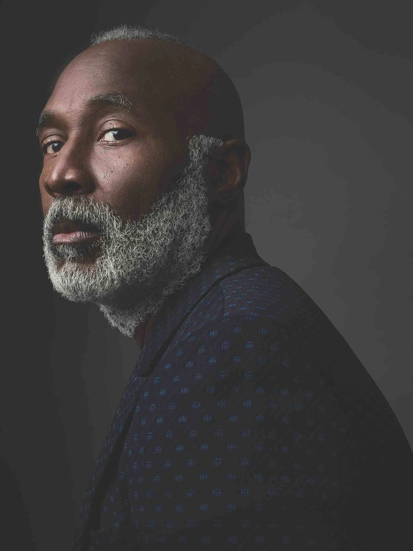 Portrait of a mature man with gray beard and hair, wearing a dark patterned jacket, against a dark background.